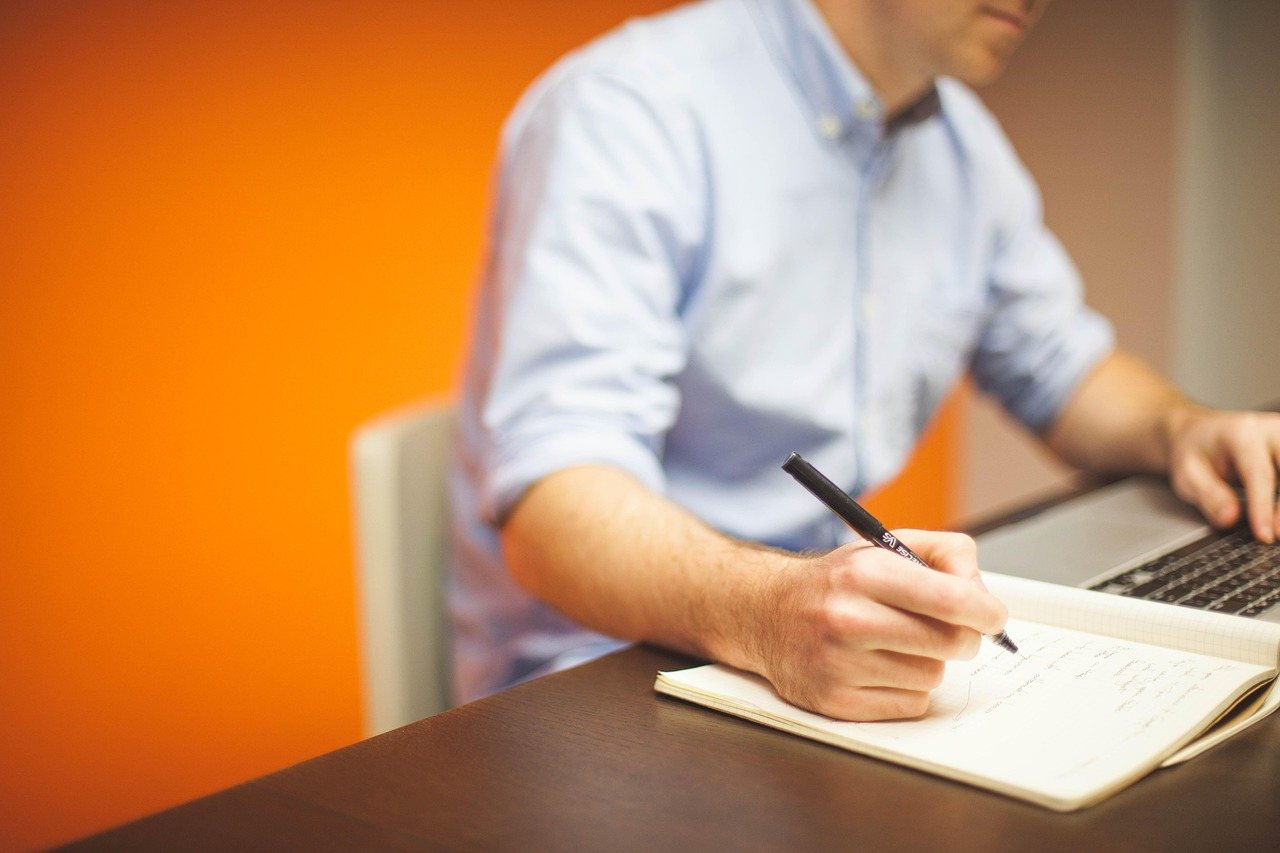 Man at a desk writing plans in a notebook next to his open laptop